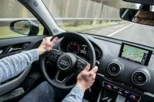 En person sitter i bil med händerna på ratten. Personens ögon där han bär glasögon syns i backspegeln