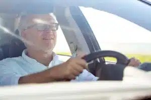 Glad bilförare i framsätet på en bil. Han har kort grått hår, svarta glasögonbågar, blå skjorta med uppkavlade ärmar.